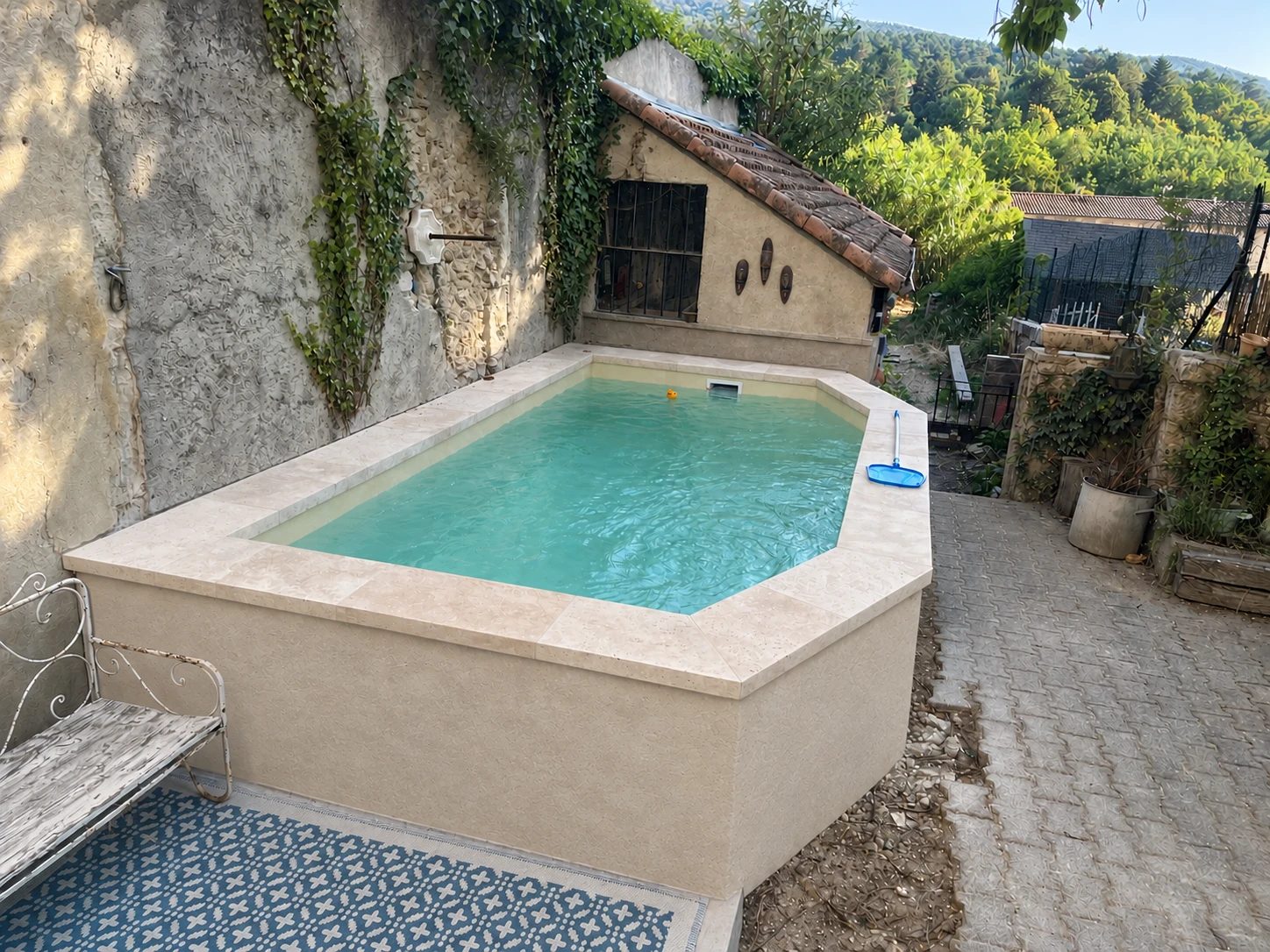Piscine surélevée maçonnée, enduit beige et margelles en pierre claire, au pied d'un mur couvert de lierre