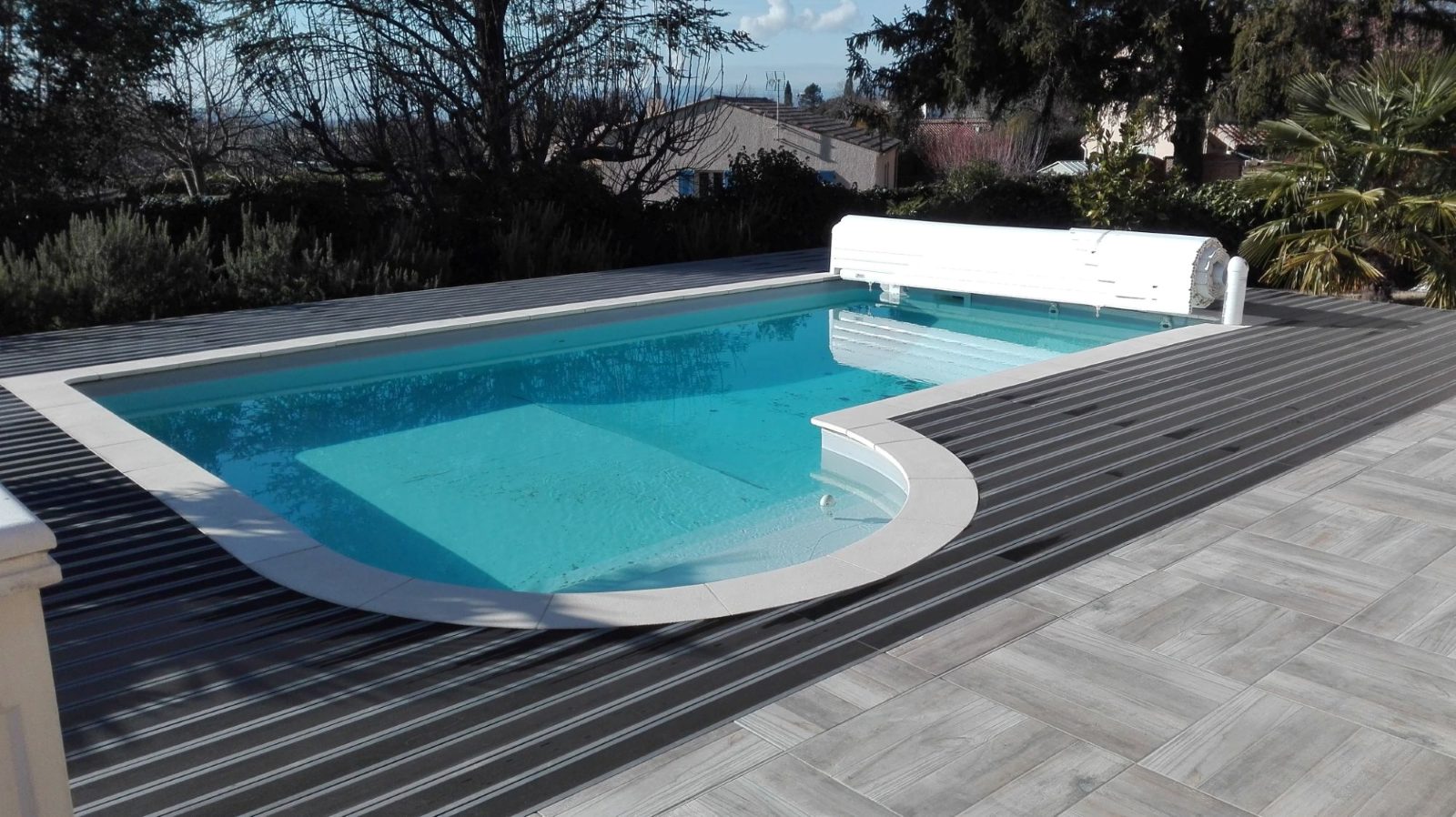 Piscine avec escalier arrondi et volet roulant, entourée d'une terrasse en lames et d'un carrelage effet bois