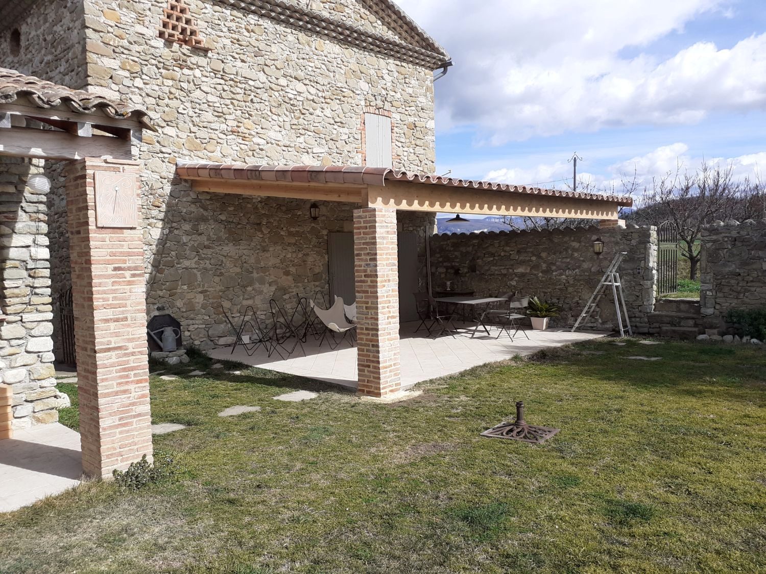 Terrasse couverte contre une maison en pierre, piliers en briques et toiture en tuiles